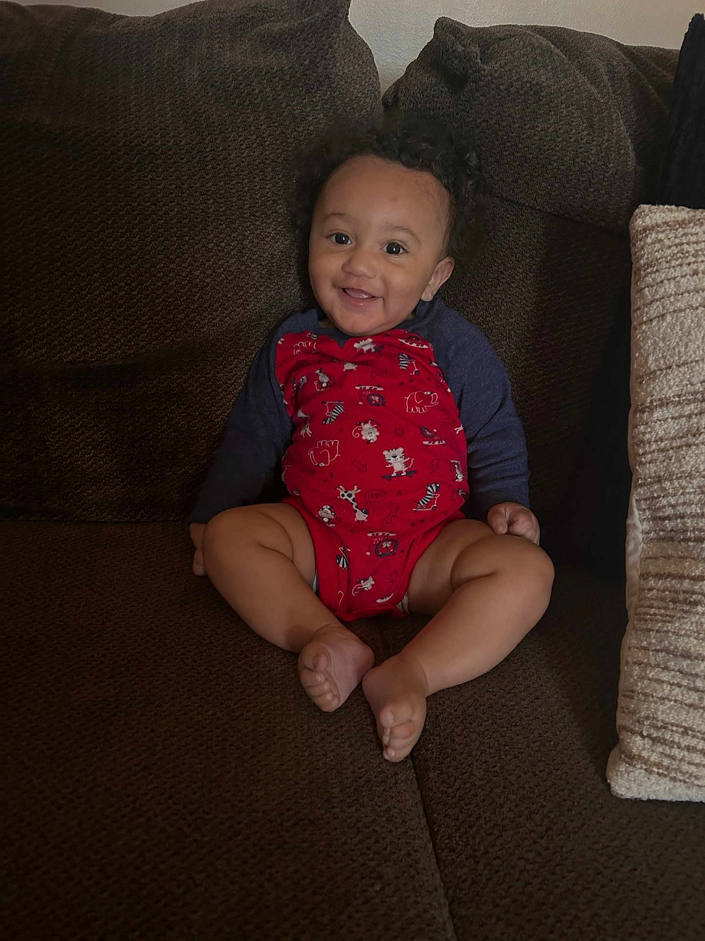 Kenzy is registered to the contest to win money with this photo: baby, child, smiling, curly_hair, red_onesie, animal_prints, couch, brown_couch, indoor, happy, sitting, barefoot, cute, infant, comfortable, plush, home, portrait, toddler, cozy