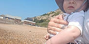 Ruben is registered to the contest to win money with this photo: baby, child, person, hat, sunhat, beach, pebble_beach, rocks, hill, sky, outdoor, summer, sunny, holding, adult, clothing, shorts, white_shirt, expression, nature