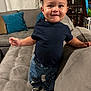 toddler, child, couch, living_room, jeans, shirt, shoes, smiling, standing, curly_hair, plush, cushions, home_interior, furniture, portrait, indoor, cute, happy, person, casual_clothing