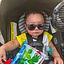 toddler, baby, car_seat, sunglasses, child, toy, pacifier, yellow_shirt, blue_shorts, book, frog, outdoor, sunlight, seatbelt, cute, serious_expression, infant, clothing, baby_toy, summer