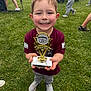 Grayson is registered to the contest to win money with this photo: child, trophy, smiling, outdoor, grass, sports, baseball, tball, happy, person, young, celebration, award, event, field, people, stroller, casual_clothing, shoes, daylight