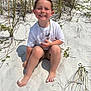 child, boy, smiling, beach, sand, barefoot, grass, sunny, outdoor, casual_clothing, white_shirt, shorts, necklace, happy, young, summer, nature, recreation, sitting, vacation