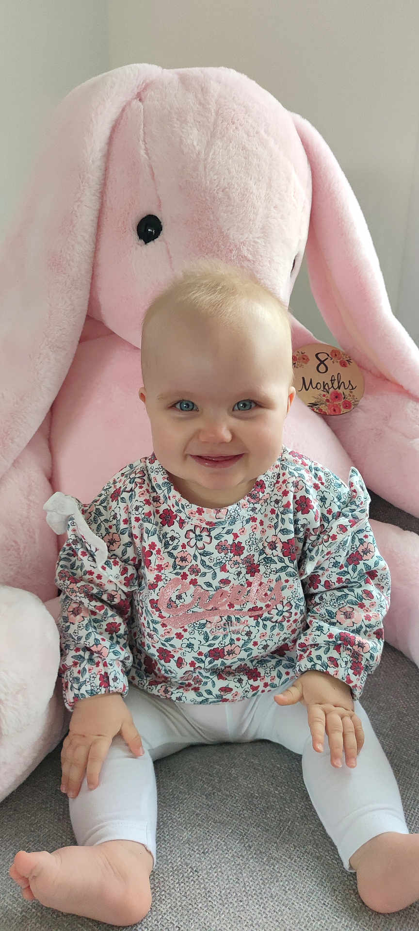 Elyne participe au concours pour gagner de l'argent avec cette photo : baby, child, smiling, sitting, plush_toy, pink, bunny, stuffed_animal, couch, clothing, floral_pattern, white_leggings, milestone, indoor, cute, happy, face, hand, feet, portrait