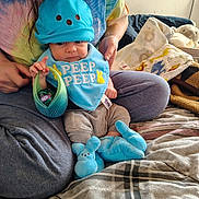 Ian is registered to the contest to win money with this photo: adult, baby, basket, bed, blanket, blue_clothing, bunny_slippers, casual_clothing, child, cozy, hat, holding, home, indoor, person, plush_toy, sitting, smile, soft_toys, tie_dye_shirt