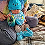 adult, baby, basket, bed, blanket, blue_clothing, bunny_slippers, casual_clothing, child, cozy, hat, holding, home, indoor, person, plush_toy, sitting, smile, soft_toys, tie_dye_shirt
