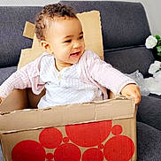 Iris a rejoint le concours — aidez-le/la à gagner de superbes lots ! toddler, child, cardboard_box, couch, smiling, curly_hair, indoor, white_clothing, pink_sweater, happy, sitting, home, playful, furniture, person, portrait, baby, cozy, relaxing, cute