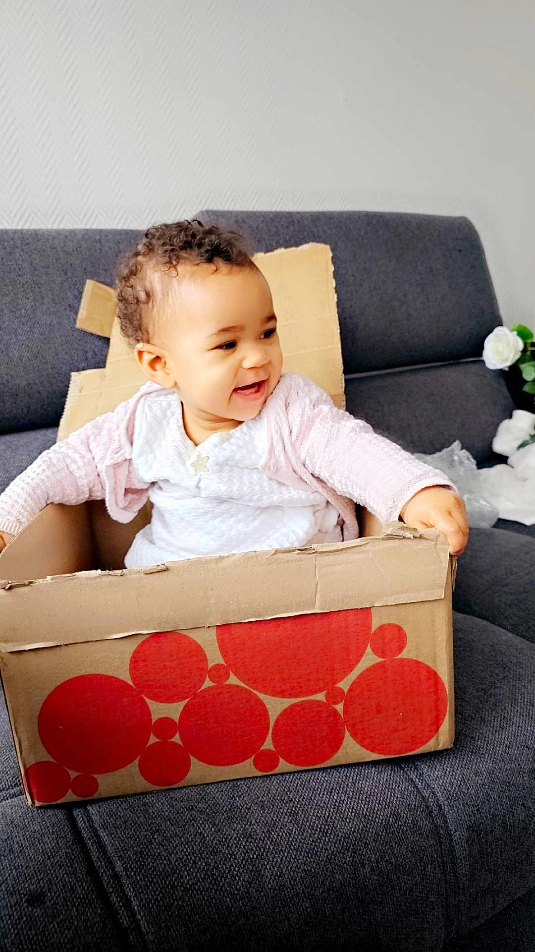 Iris a rejoint le concours — aidez-le/la à gagner de superbes lots ! toddler, child, cardboard_box, couch, smiling, curly_hair, indoor, white_clothing, pink_sweater, happy, sitting, home, playful, furniture, person, portrait, baby, cozy, relaxing, cute