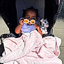 baby, stroller, blanket, plush_toy, sunglasses, flower_shaped, orange_lenses, pink_blanket, soft_texture, child, outdoor, sunlight, curious, infant, toy, foot, clothing, seat, shade, playful