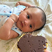 Ibrahim joined the competition — help win amazing prizes! baby, infant, face, hand, chocolate, biscuit, food, blanket, bed, curious, lying_down, person, skin, expression, closeup, focus, comfort, indoor, soft_lighting, nourishment