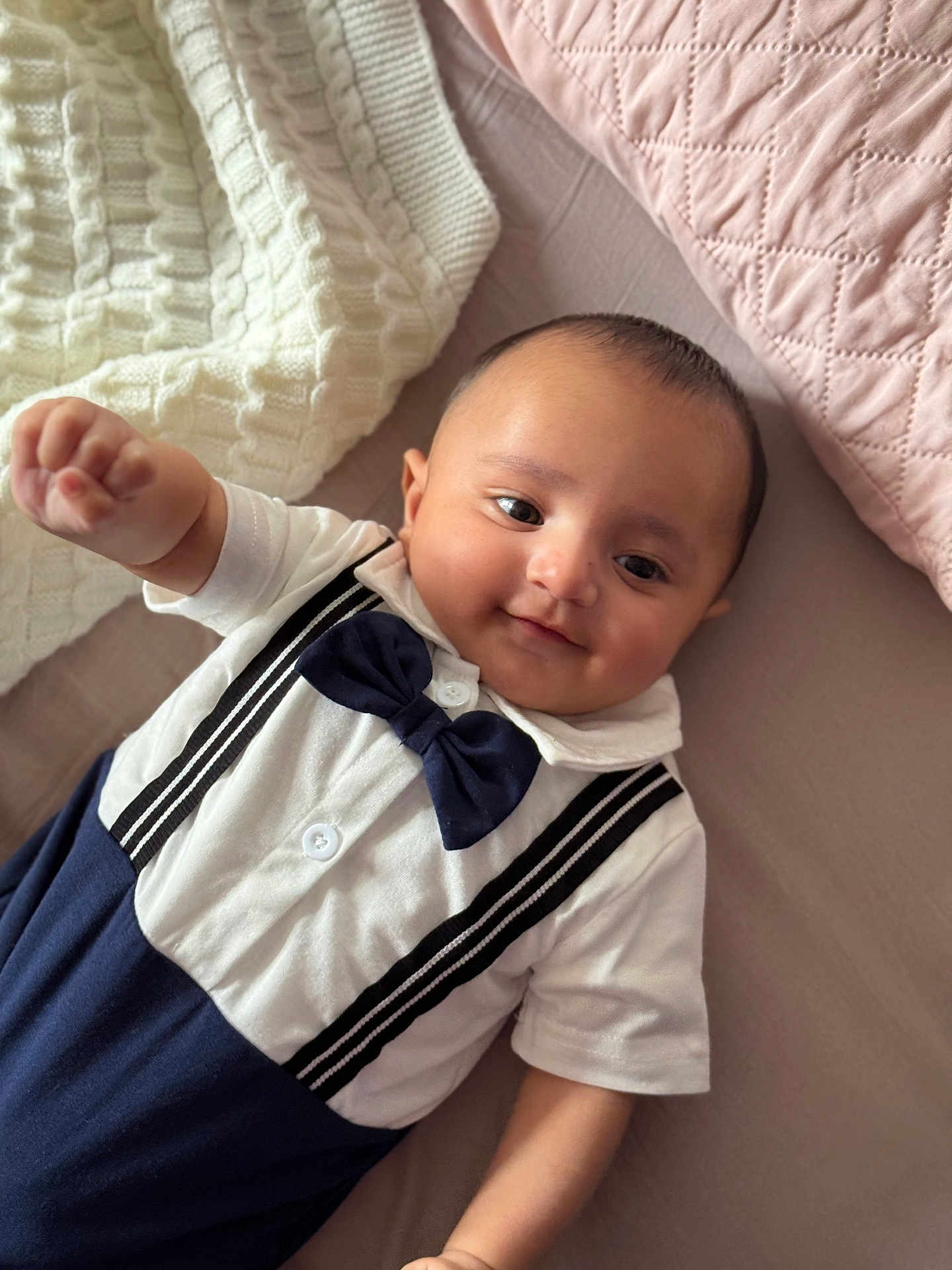 Ibrahim is registered to the contest to win money with this photo: baby, infant, smiling, bow_tie, suspenders, white_shirt, navy_pants, bed, blanket, pillow, cute, portrait, child, person, indoors, soft_lighting, happy, lying_down, closeup, young