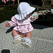 Lilah is registered to the contest to win money with this photo: baby, child, hat, sun_hat, pacifier, pink_dress, white_shoes, socks, outdoor, grass, bicycle, mat, shadow, person, sitting, toy, summer, sunlight, playtime, relaxation