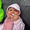 baby, child, pink_hoodie, bib, snack, smile, face, happy, cushion, gray_background, hand, cheeks, eyes, indoors, cozy, clothing, person, cute, infant, munching