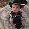 baby, child, cowboy_hat, portrait, sitting, infant, serious_expression, wide_eyes, denim_overalls, plaid_shirt, crib, baby_seat, cushion, blanket, bedroom, indoors, patterned_fabric, cowboy_costume, cute, photography