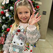 Velouria is registered to the contest to win money with this photo: child, girl, blonde_hair, pink_bows, waving_hand, smiling, festive_sweater, tinsel, bells, hello_kitty, christmas_tree, ornaments, holiday_decor, indoor, portrait, happy, tiled_floor, home, holiday_season, cute