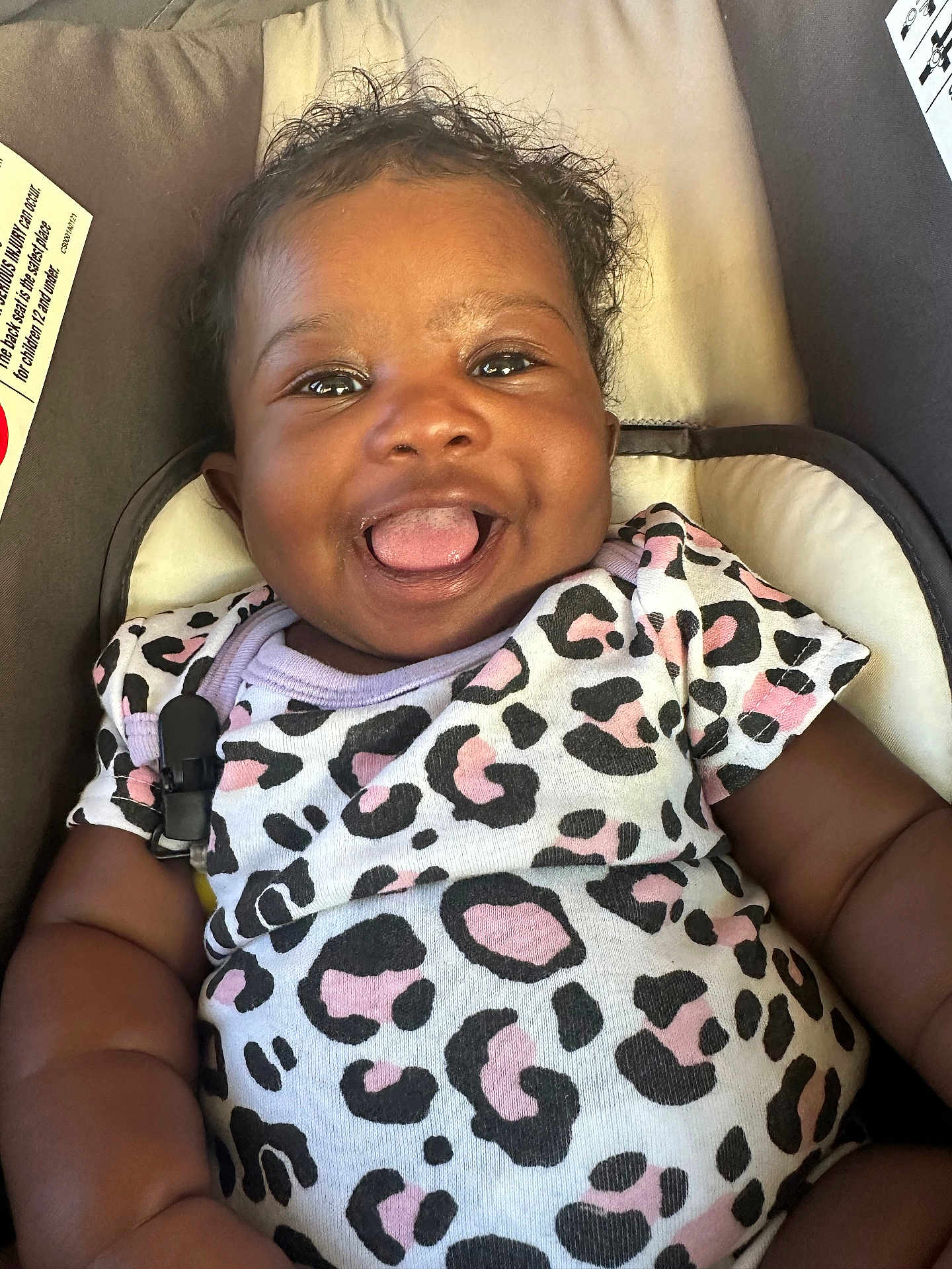 Malina is registered to the contest to win money with this photo: baby, smiling, tongue_out, leopard_print, onesie, curly_hair, infant, seat, cushion, indoor, cute, happy, child, portrait, closeup, person, playful, clothing, infancy, babywear