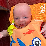 Wesley is registered to the contest to win money with this photo: baby, baby_accessory, baby_girl_or_boy, blue, blue_eyes, child, closeup, clothing, cute, face, fish_pattern, floatation_vest, happy, indoor, infant, orange, portrait, red_background, shark_design, smile