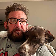Willow Mae is registered to the contest to win money with this photo: affection, beard, casual_clothing, closeup, companion, couch, cozy, dog, face, furniture, glasses, gray_shirt, head, human, indoor, man, pet, portrait, relaxed, snuggling