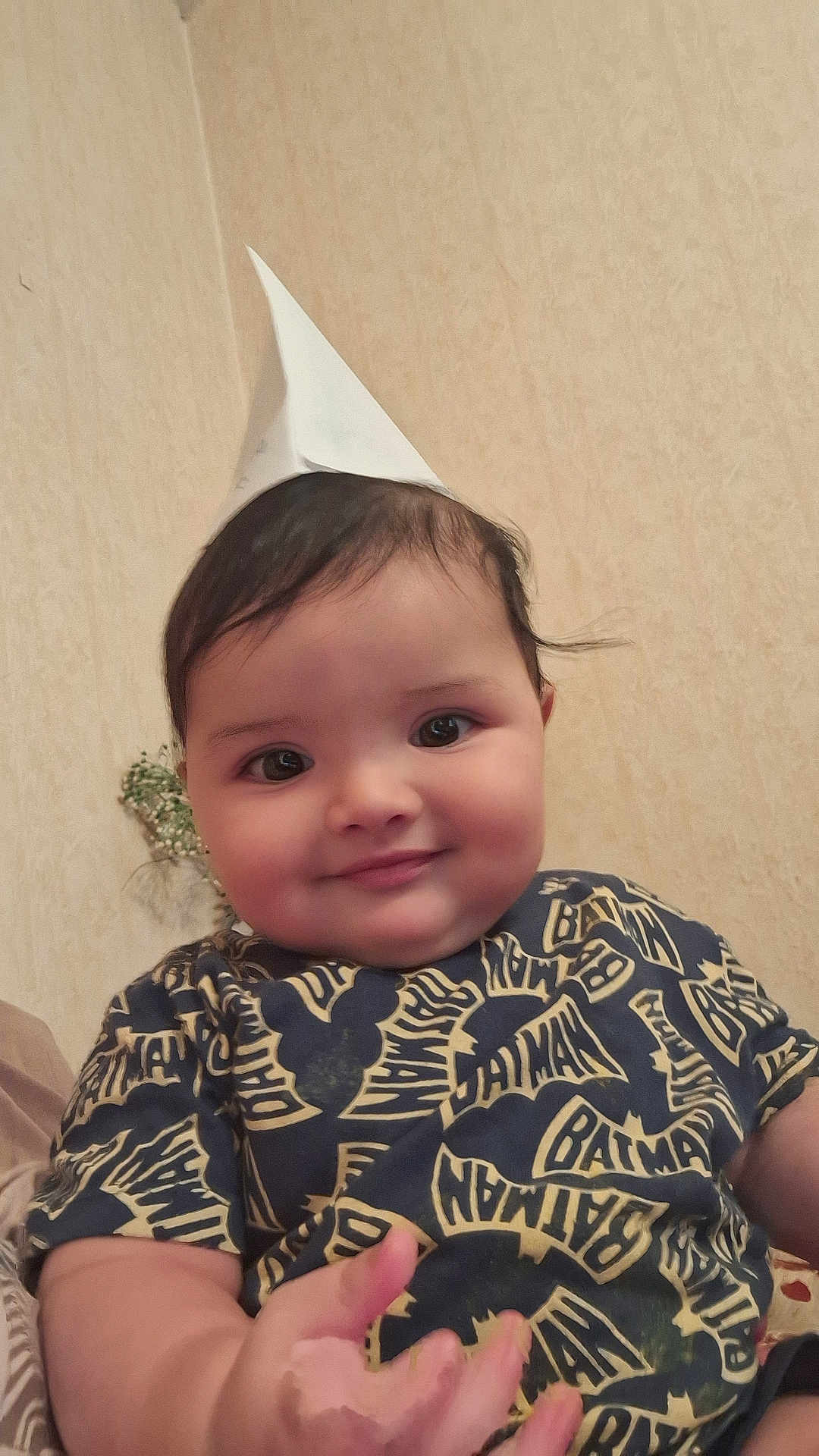 Younes is registered to the contest to win money with this photo: baby, child, smiling, paper_hat, batman_shirt, indoor, person, cute, portrait, face, cheeks, happy, young_child, hand, clothing, dark_hair, seated, wallpaper, infant, playful
