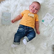 Liam a rejoint le concours — aidez-le/la à gagner de superbes lots ! baby, infant, child, smile, orange_shirt, jeans, white_rug, wooden_floor, milestone_cards, lying_down, cute, fluffy, flooring, casual_clothing, indoor, happy, portrait, cheeky, young_child, bright_colors