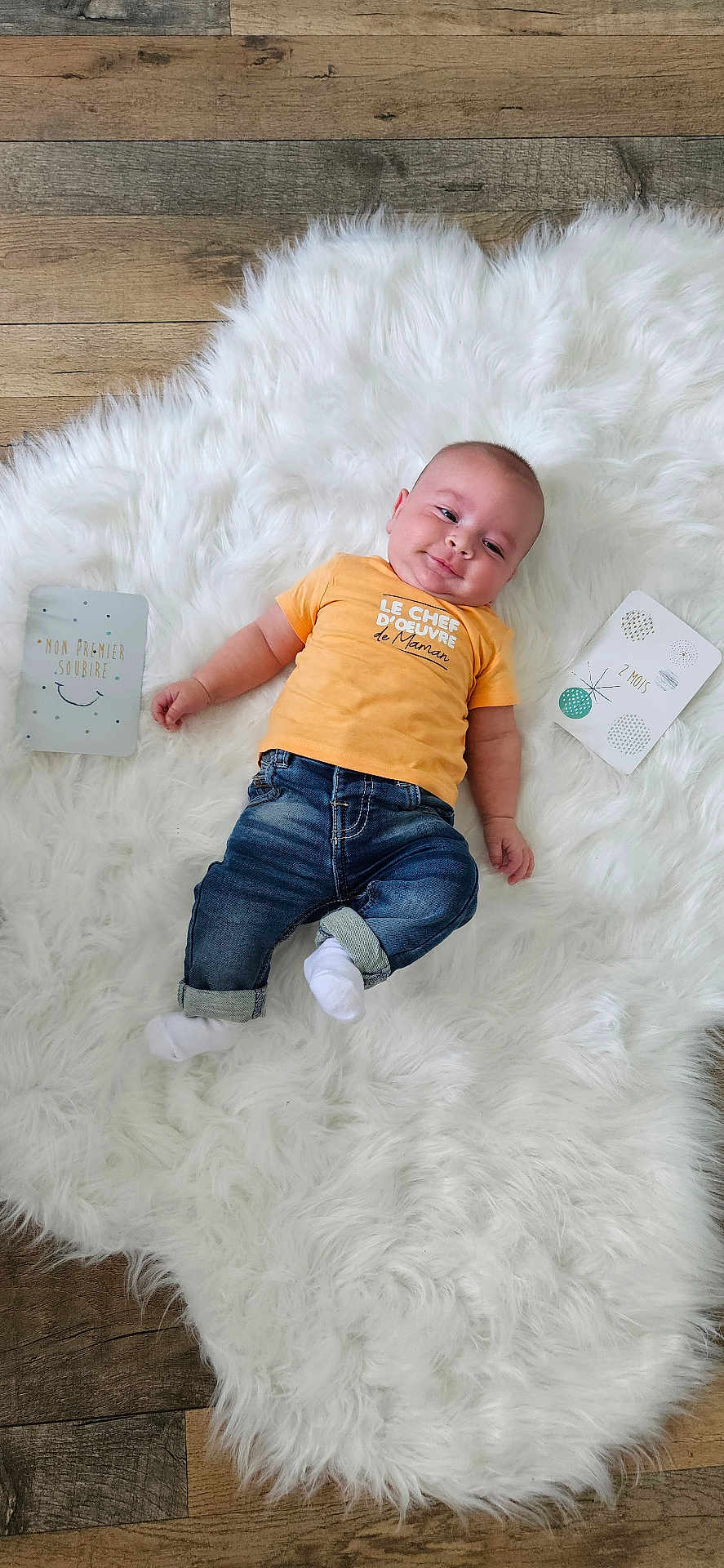 Liam a rejoint le concours — aidez-le/la à gagner de superbes lots ! baby, infant, child, smile, orange_shirt, jeans, white_rug, wooden_floor, milestone_cards, lying_down, cute, fluffy, flooring, casual_clothing, indoor, happy, portrait, cheeky, young_child, bright_colors
