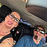 woman, man, dog, car_interior, selfie, sunglasses, cap, seatbelt, tattoo, earrings, necklace, black_dog, smiling, tongue_out, happy, daylight, window, headrest, casual_clothing, road