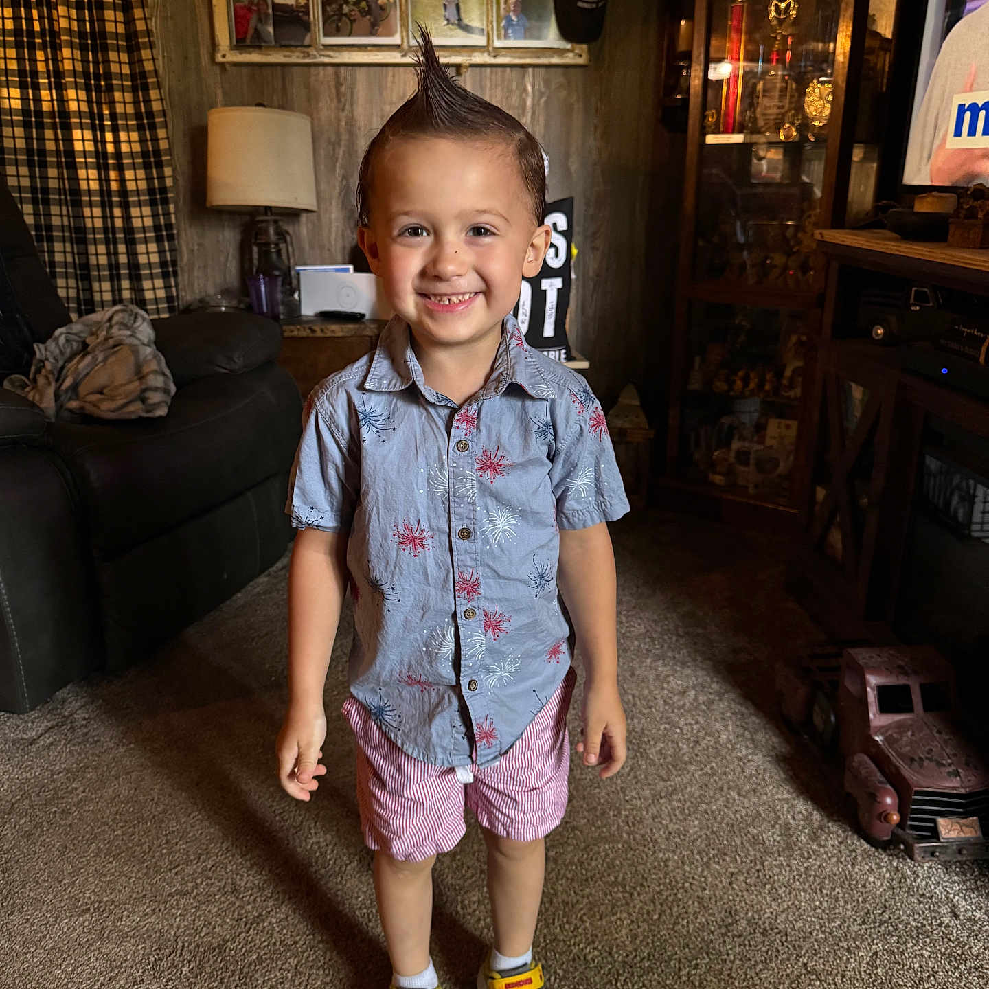 Dallas is registered to the contest to win money with this photo: boy, cabinet, carpet, child, couch, curtains, family_photos, happy, indoors, lamp, living_room, mohawk, person, shirt, shorts, smiling, sneakers, toy_truck, trophies, wood_paneling