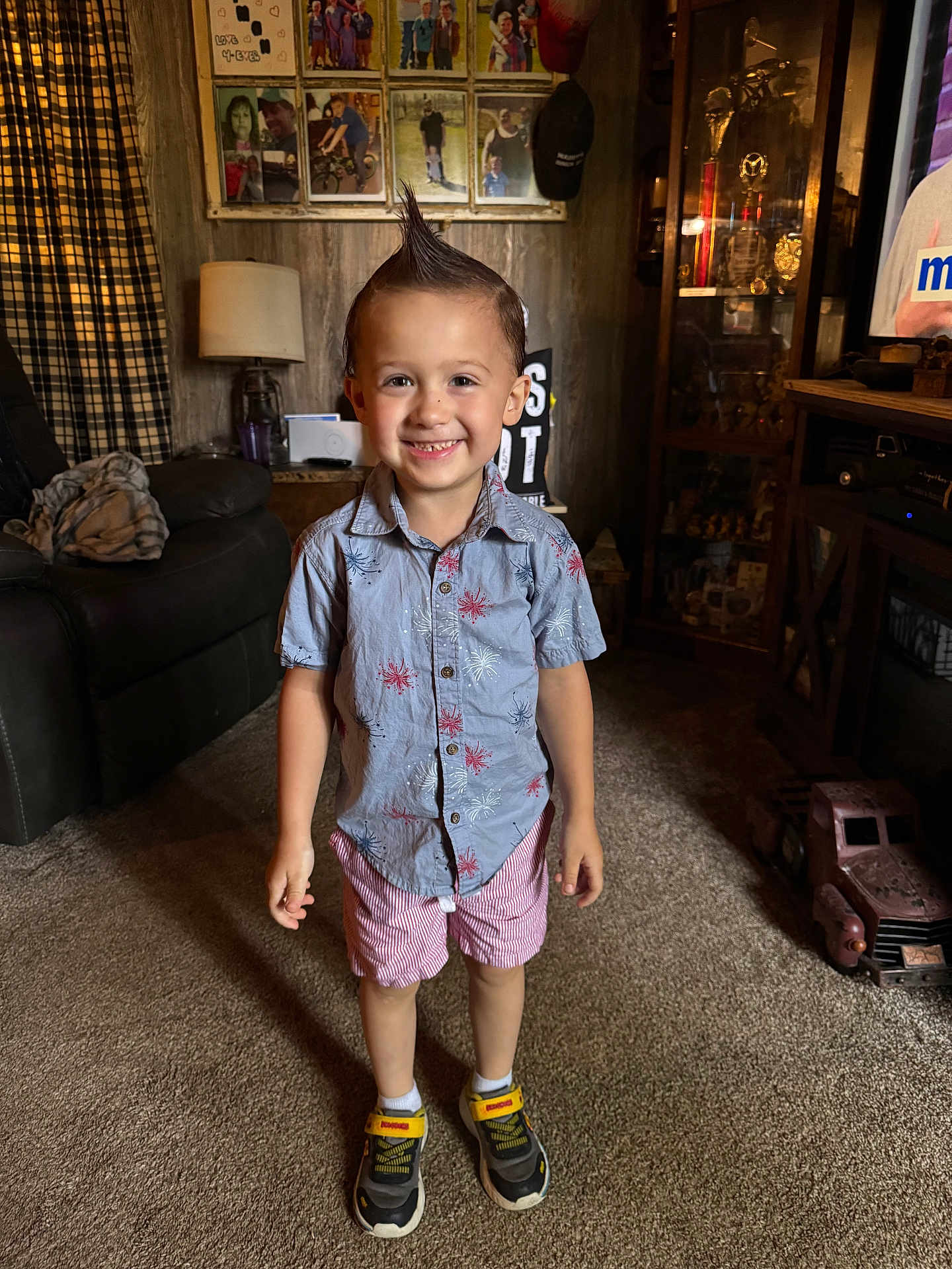 Dallas is registered to the contest to win money with this photo: child, boy, smiling, mohawk, shirt, shorts, sneakers, carpet, living_room, toy_truck, trophies, family_photos, lamp, couch, curtains, wood_paneling, cabinet, happy, indoors, person
