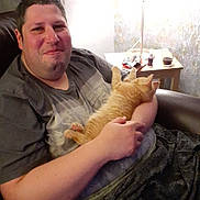 Walter is registered to the contest to win money with this photo: man, cat, kitten, smiling, sitting, chair, lamp, table, drink, snacks, indoor, cozy, relaxed, casual_clothing, home, pet, orange_cat, comfort, happy, portrait