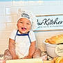 baby, chef_hat, apron, flour, kitchen, baking, bread, croissant, rolling_pin, smiling, happy, infant, countertop, white_tiles, sign, cooking, cute, messy, child, portrait