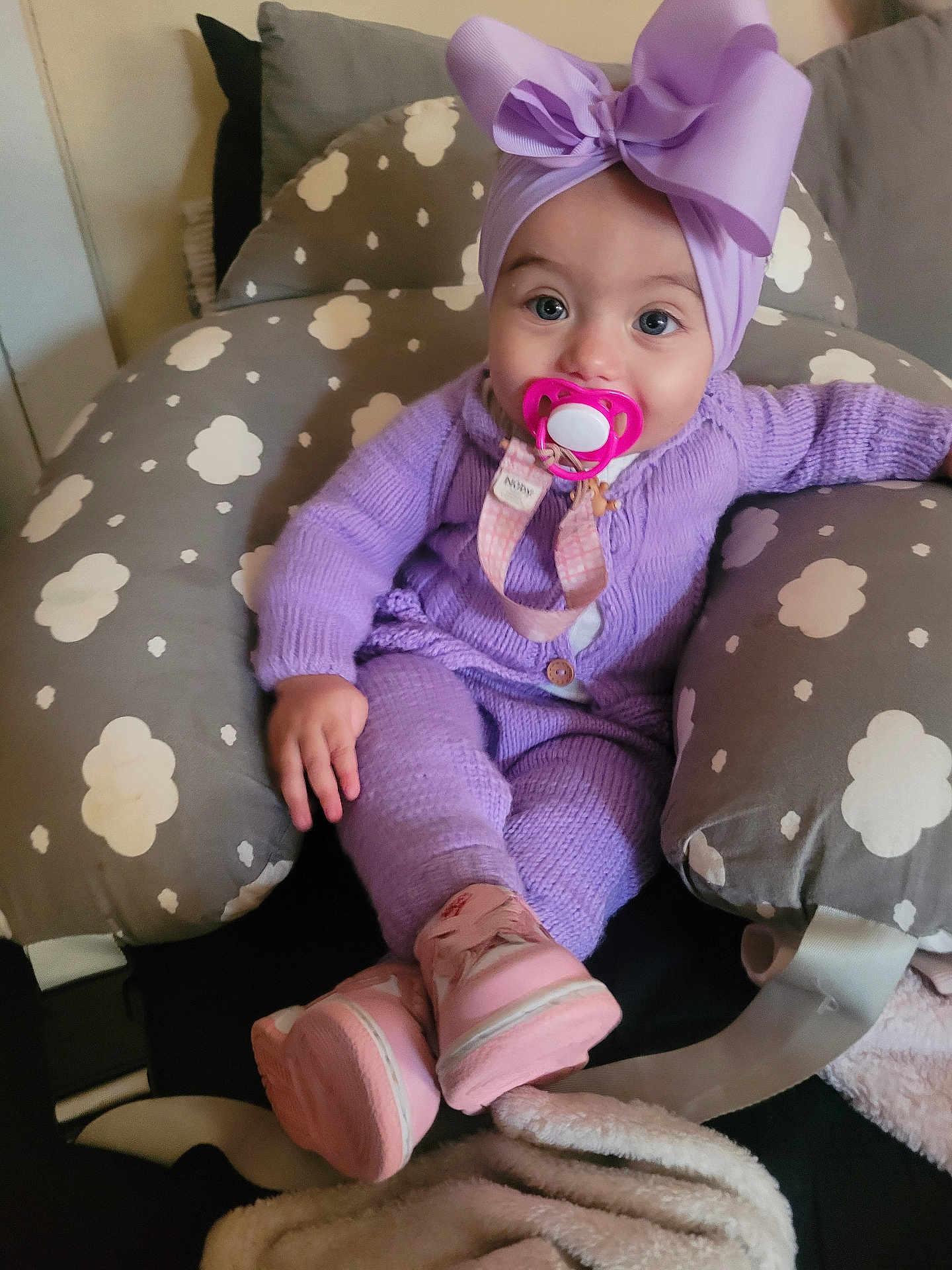 Suzanna is registered to the contest to win money with this photo: baby, infant, pacifier, purple_outfit, headwrap, bow, pink_boots, baby_seat, cloud_pattern, knitwear, cozy, cute, indoors, child, sitting, soft_toys, blanket, comfort, portrait, young_child
