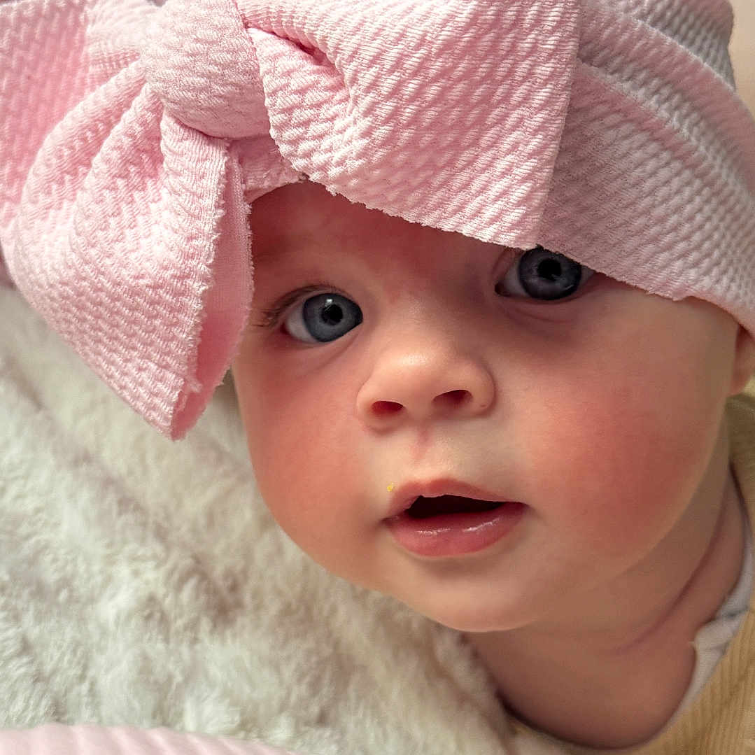 Meivė is registered to the contest to win money with this photo: baby, blanket, blue_eyes, child, closeup, cozy, curious, cute, expression, face, head, headwear, indoor, infant, person, pink_bow, portrait, skin, soft_texture, young
