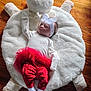 baby, infant, sleeping, headband, bow, red_pants, white_top, pacifier, plush_cushion, bear_shape, wooden_floor, cozy, cute, toddler, child, indoors, soft_texture, resting, peaceful, adorable