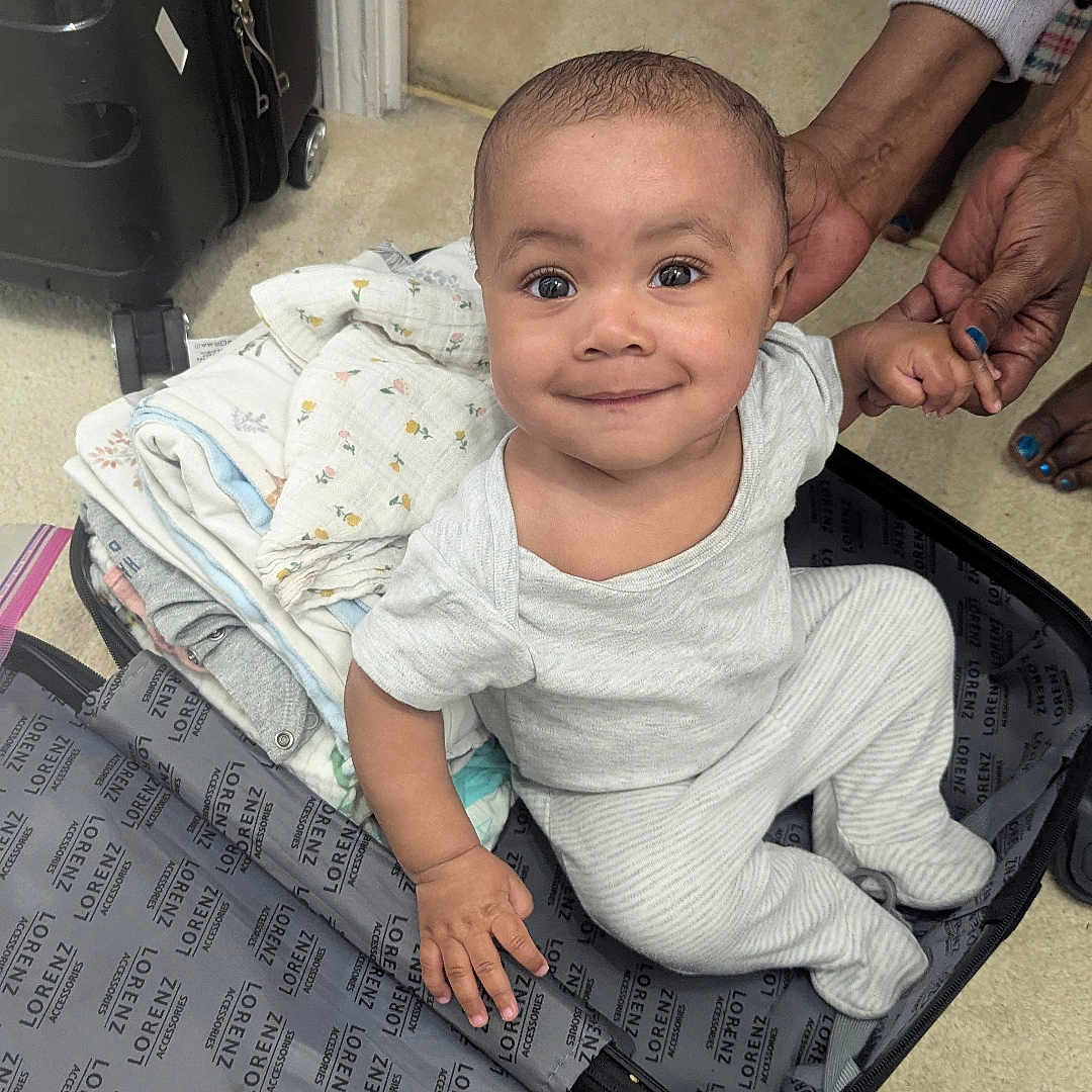 Demari is registered to the contest to win money with this photo: adult_hand, baby, beige_floor, child, clothing, content, curious, floor, folded_clothes, hand, holding_hands, indoor, luggage, open_suitcase, pajamas, person, smile, striped_clothing, suitcase, travel