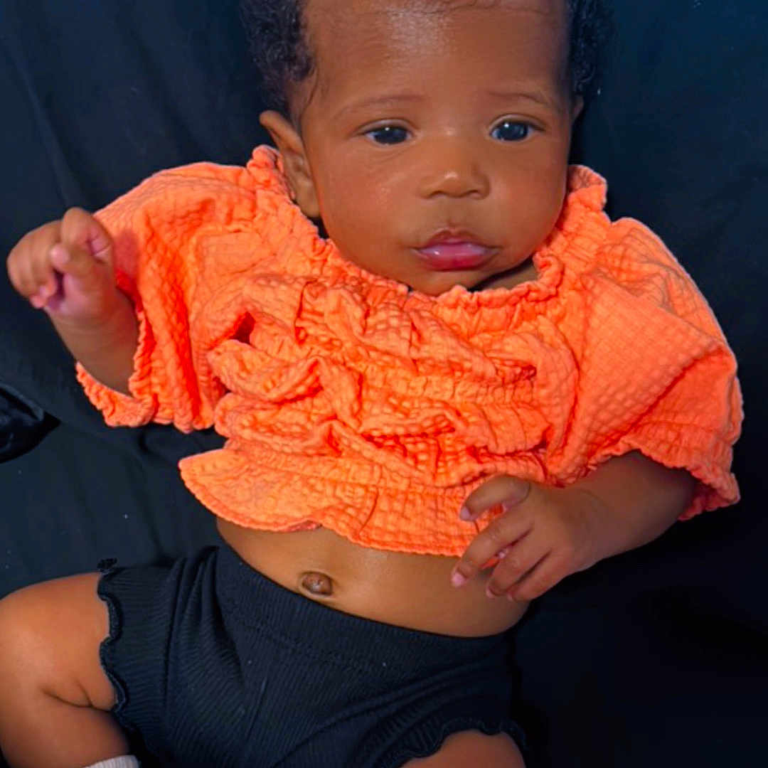 Brigette is registered to the contest to win money with this photo: adorable, baby, black_shorts, bow, child, closeup, curly_hair, cute, dark_background, face, feet, hands, infant, lying_down, newborn, orange_clothing, portrait, smiling, socks, striped_bow