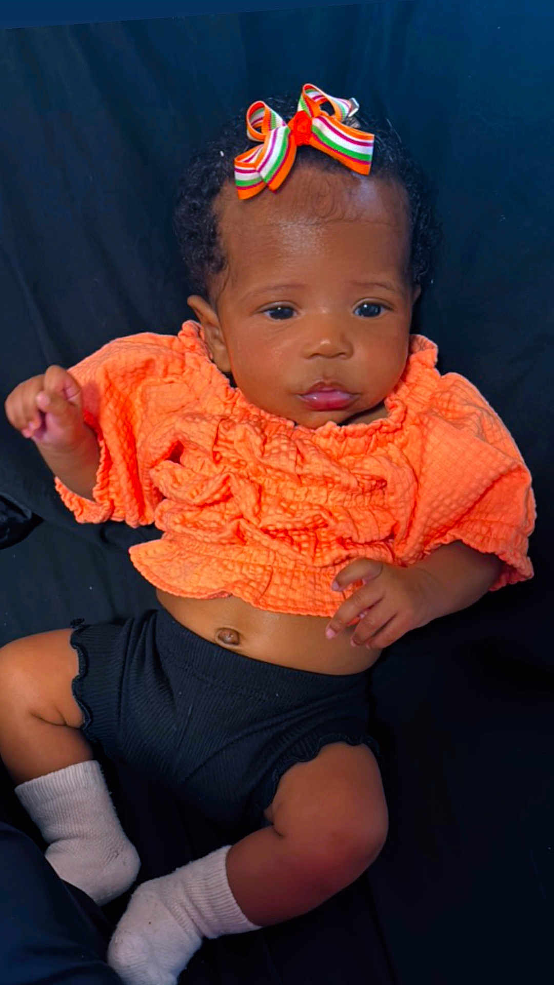 Brigette is registered to the contest to win money with this photo: baby, infant, child, orange_clothing, black_shorts, bow, striped_bow, socks, lying_down, portrait, cute, dark_background, smiling, face, hands, feet, newborn, curly_hair, adorable, closeup