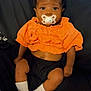 Brigette is registered to the contest to win money with this photo: baby, infant, pacifier, bow, orange_clothing, black_shorts, white_socks, cute, child, portrait, indoors, sitting, looking_at_camera, relaxed, dark_background, soft_fabric, small_person, adorable, young_child, casual_clothing