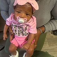 Brigette joined the competition — help win amazing prizes! baby, infant, child, person, pacifier, pink_clothing, headband, shoes, adult_hands, holding, indoor, flooring, sitting, cute, small, accessory, portrait, human, casual, family