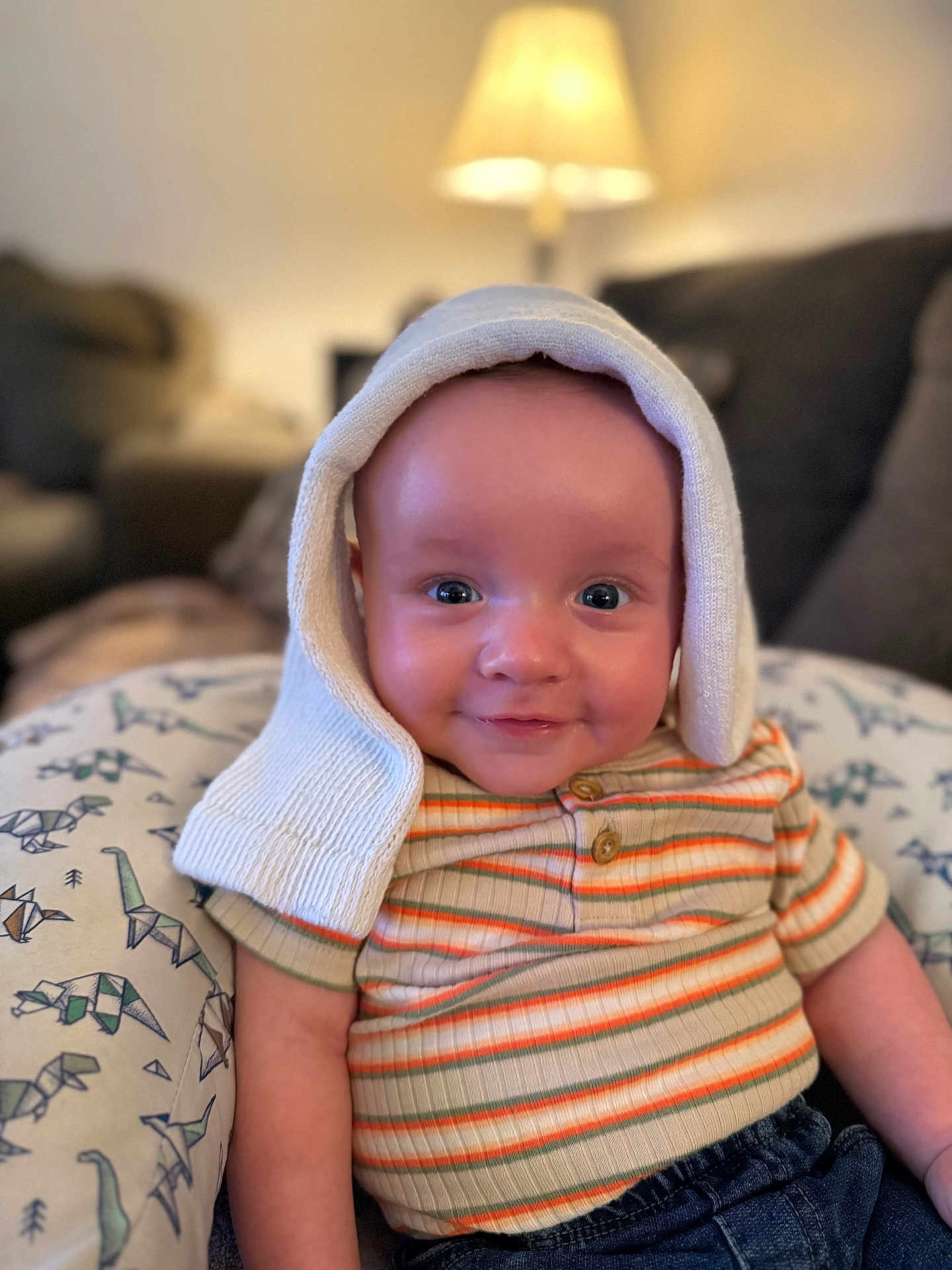 Jameson is registered to the contest to win money with this photo: baby, infant, smile, striped_shirt, hat, knit, dinosaur_pattern, pillow, indoor, lamp, cozy, cute, child, portrait, seated, denim, happy, face, eyes, warm_lighting