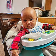 Karter joined the competition — help win amazing prizes! baby, baby_seat, bib, blanket, cabinet, carpet, child, curious, expression, face, furniture, home, indoor, infant, kitchen, person, plaid_clothing, table, toy, wooden_chair