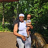 Karter is registered to the contest to win money with this photo: baby, casual_clothing, child, curious, daytime, dinosaur_statue, expression, greenery, headband, holding, nature, outdoor, park, person, playground, sculpture, shadow, sunlight, trees, woman