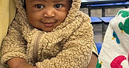 Karter is registered to the contest to win money with this photo: baby, background_person, bench, cafeteria, child, colorful_cloth, cute, face, food_wrapper, fuzzy_clothing, hand, holding, indoor, infant, person, portrait, smile, table, teddy_bear_hoodie, warm_clothing
