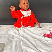 Alfredinho participe au concours pour gagner de l'argent avec cette photo : baby, child, red_onesie, bed, blanket, headboard, indoor, portrait, sitting, curious, infant, white_blanket, baby_bib, expression, face, looking_away, clothing, young_child, home, casual
