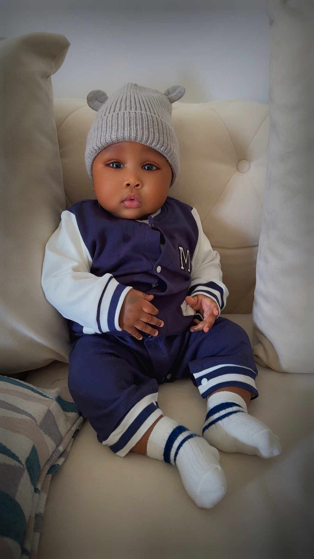 Lucas is registered to the contest to win money with this photo: baby, child, infant, couch, hat, knit_hat, bear_ears, navy_jacket, white_sleeves, socks, sitting, pillow, beige, indoor, portrait, cute, expression, hands, feet, comfort
