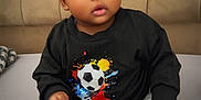 Lucas is registered to the contest to win money with this photo: baby, child, infant, sitting, bed, black_sweatshirt, soccer_ball, curious, indoors, headboard, plush, feet, hands, clothing, portrait, young, cute, expression, casual, comfortable