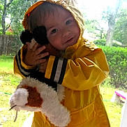 Samuel is registered to the contest to win money with this photo: toddler, child, yellow_raincoat, stuffed_animal, hugging, indoor, window, outdoor, greenery, raincoat_hood, plush_toy, cute, portrait, smiling, young_child, playing, cozy, childhood, happy, soft_light