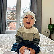 Nathanael participe au concours pour gagner de l'argent avec cette photo : baby, child, smiling, hat, sweater, white_shoes, indoor, window, curtains, plant, bed, blanket, happy, person, portrait, cozy, cute, sitting, home, natural_light
