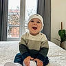 baby, child, smiling, hat, sweater, white_shoes, indoor, window, curtains, plant, bed, blanket, happy, person, portrait, cozy, cute, sitting, home, natural_light