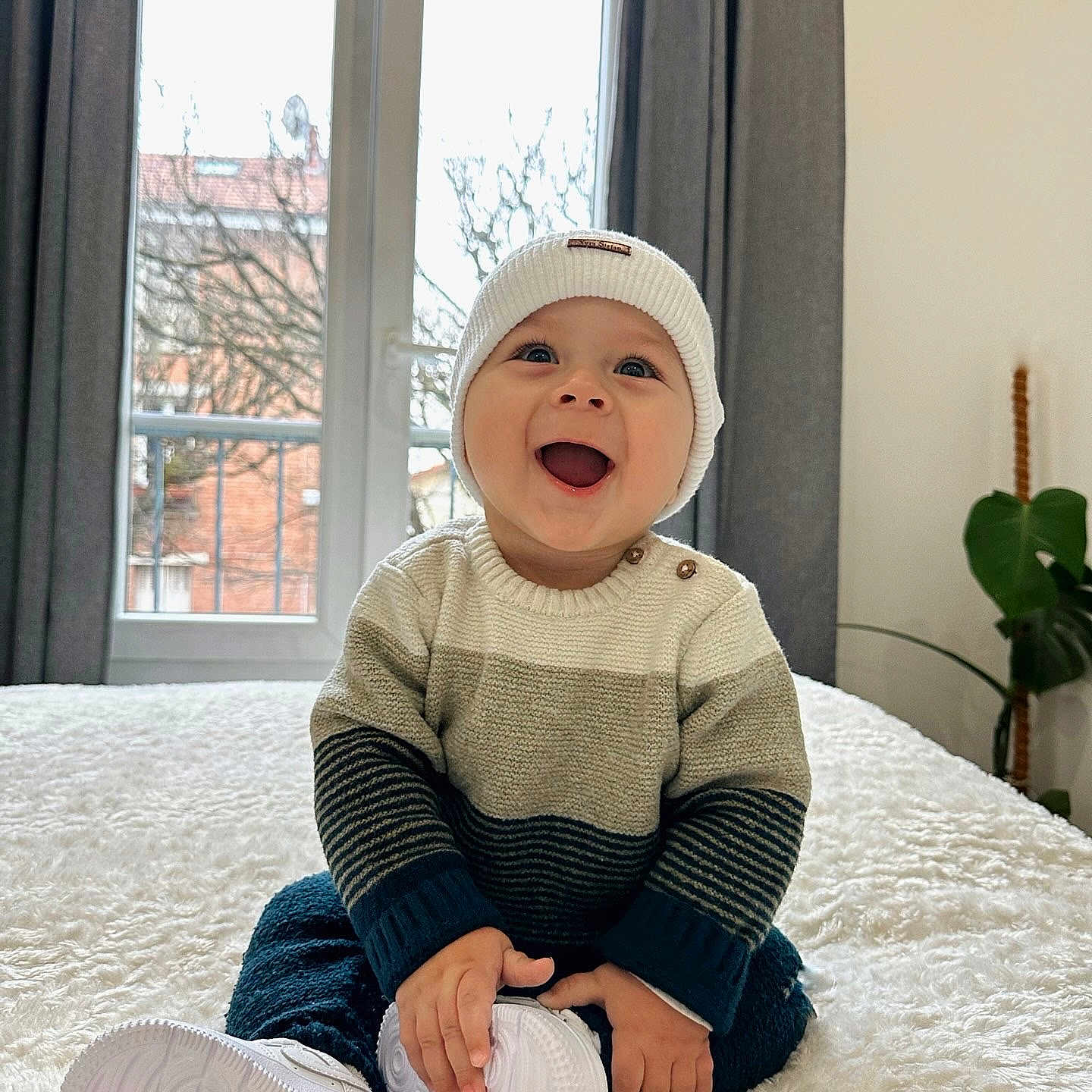 Nathanael participe au concours pour gagner de l'argent avec cette photo : baby, child, smiling, hat, sweater, white_shoes, indoor, window, curtains, plant, bed, blanket, happy, person, portrait, cozy, cute, sitting, home, natural_light
