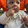 baby, child, person, face, hand, finger, clothing, onesie, orange_shirt, outdoor, grass, fence, car, seated, curious, closeup, portrait, expression, skin, casual