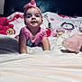 baby, pink_headband, pink_shirt, stuffed_animals, bed, blanket, floral_pattern, teddy_bear, child, cute, smiling, indoors, portrait, infant, toy, cozy, happy, lying_down, headband, soft_toys